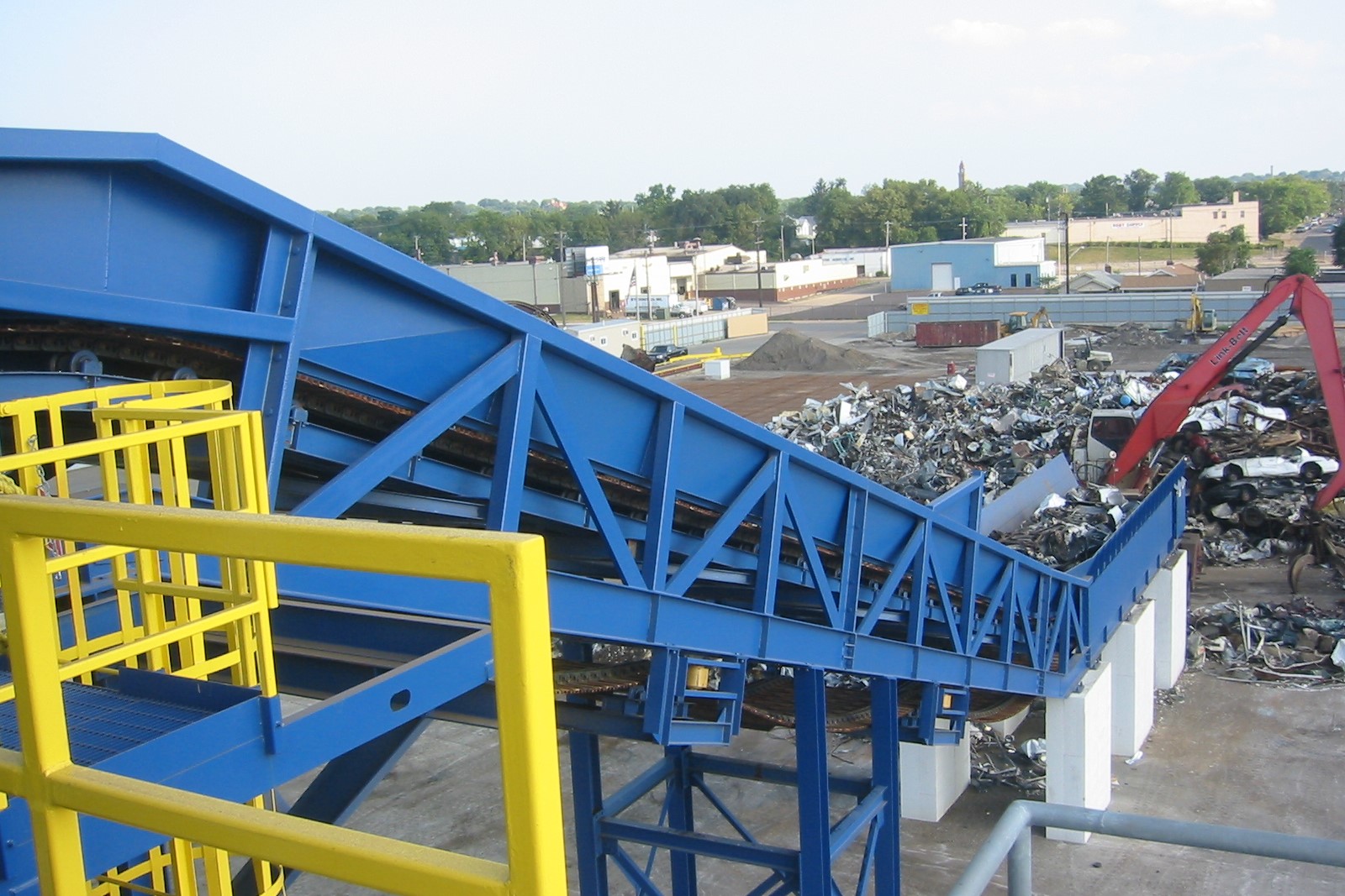 5eb0697b1e92213d60ec40bc_IMG_4664 (2) A blue industrial conveyor belt structure at an industrial metal recycling or scrap yard, transporting materials over a large pile of metal debris, with buildings and trees visible in the background.