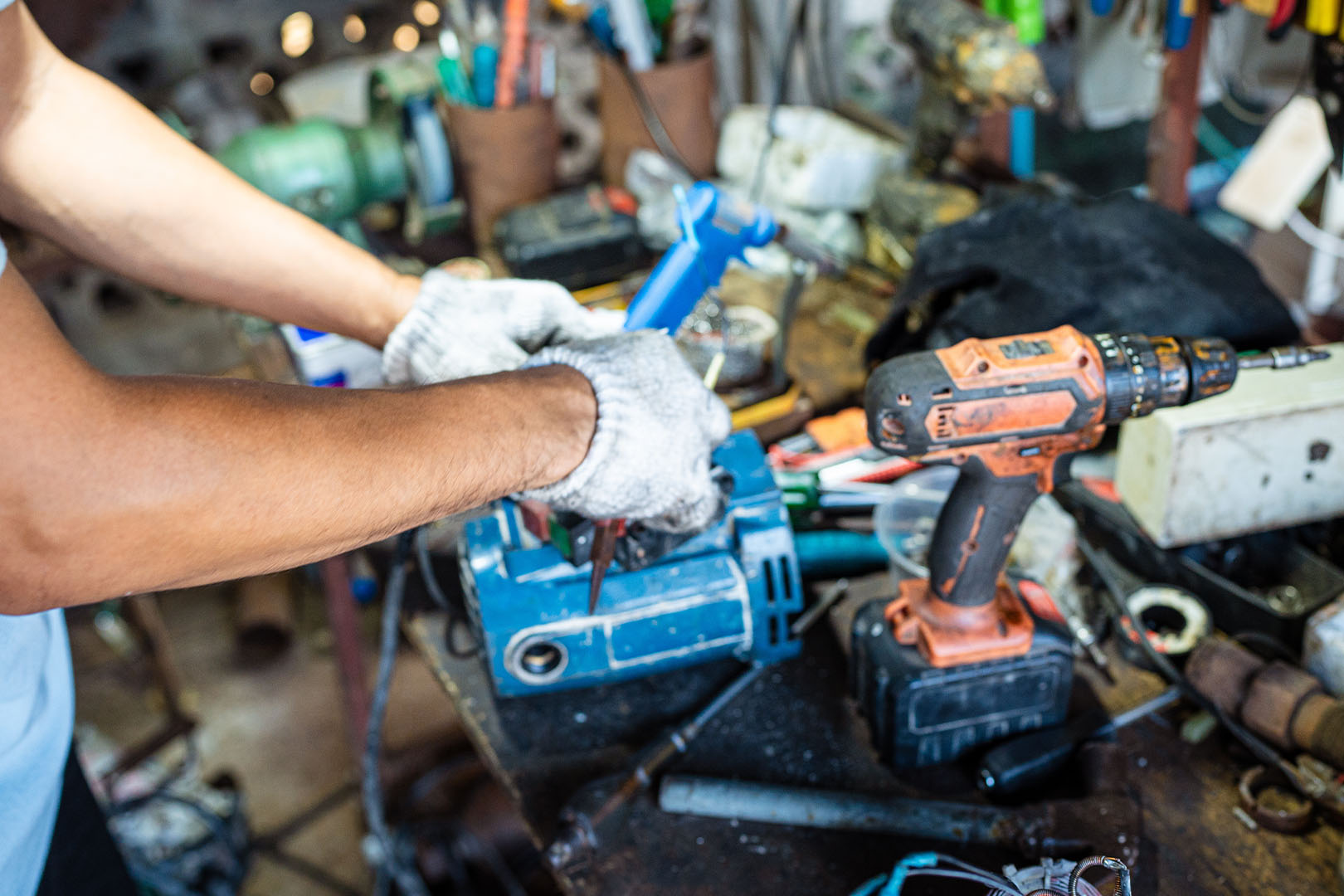 tools-spot A person wearing gray gloves works with tools on a cluttered workbench, focused on Industrial Tool Repair. An orange cordless drill and a blue power tool are visible among various scattered equipment and parts.